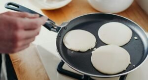 Close-up of pancakes cooking in a pan on a gas stove with utensils nearby.
