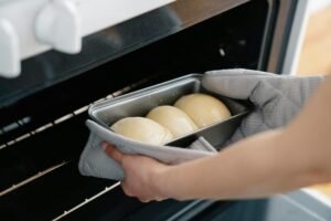 Person placing dough buns in oven, wearing gloves and using baking tray. Captured mid-preparation in a kitchen setting.