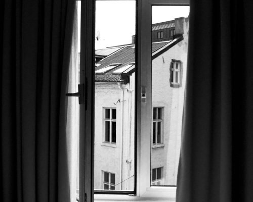 A monochrome view of an exterior building through an open window with curtains.